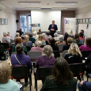 Starszy mężczyzna w garniturze zwrócony przodem do publiczności prowadzi spotkanie.