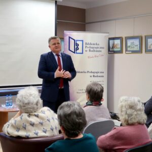 Starszy mężczyzna w garniturze zwrócony przodem do publiczności prowadzi spotkanie.