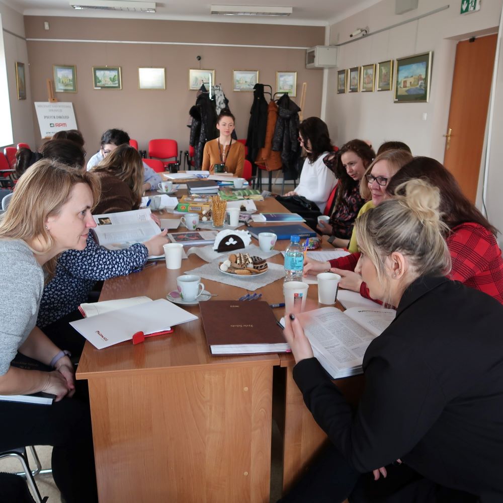 Grupa pań siedzących przy stole konferencyjnym uważnie słucha prelegentki.