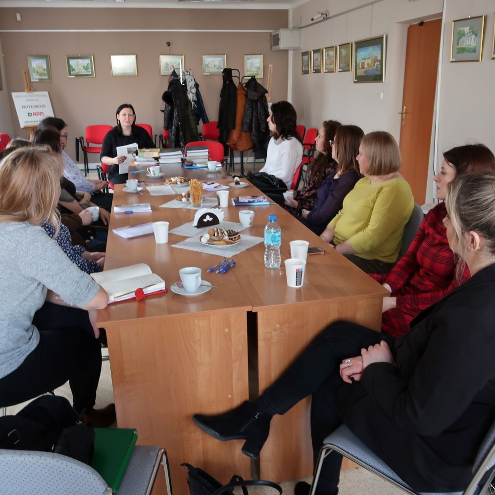 Grupa pań siedzących przy stole konferencyjnym uważnie słucha prelegentki.
