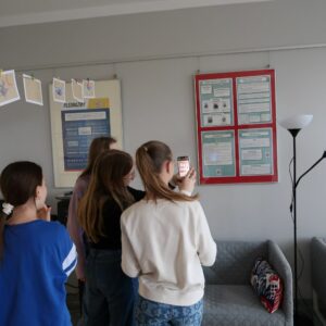 obchodody-Międzynarodowego-Dnia-Jezyka-Ojczystego-uczniowie-PSP im. Jana-Kochanowskiego-w-Zakrzewie-5 Grupa uczennic robi zdjęcie smartfonem tablicy informacyjnej zawieszonej na ścianie.