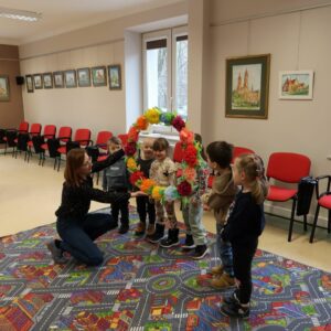 Dzieci pozują do zdjęcia stojąc w szeregu, pani bibliotekarka podaje im wieniec stworzony z papierowych kwiatów.