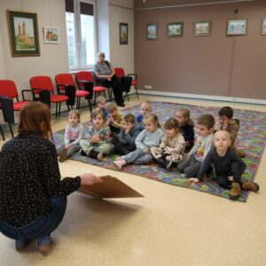 Grupa dzieci z zaciekawieniem słucha pani bibliotekarki opowiadającej o zbiorach bibliotecznych