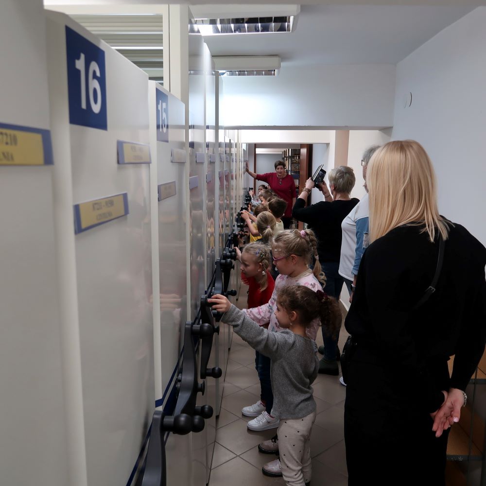 dzien-przytulania-bibliotekarza-2 Dzieci w magazynie bibliotecznym przesuwają regały na szynach