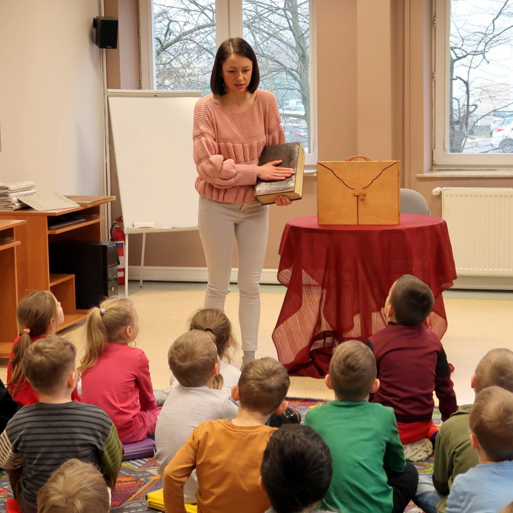 Dzieci siedząc na podłodze oglądają książkę pokazywaną przez bibliotekarkę