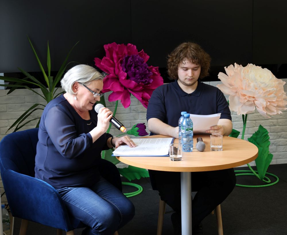 konkurs-tworczosc-literacka-5 Kobieta z mikrofonem w ręce odczytuje tekst zapisany na kartce obok młody mężczyzna przegląda kartki trzymane w ręce.