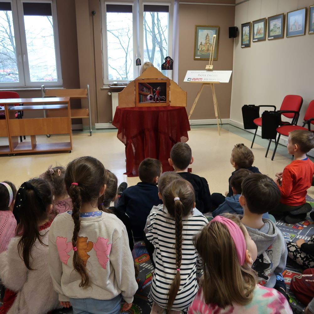 Dzieci siedząc na dywanie słuchają opowieści wpatrzone w teatrzyk Kamishibai.