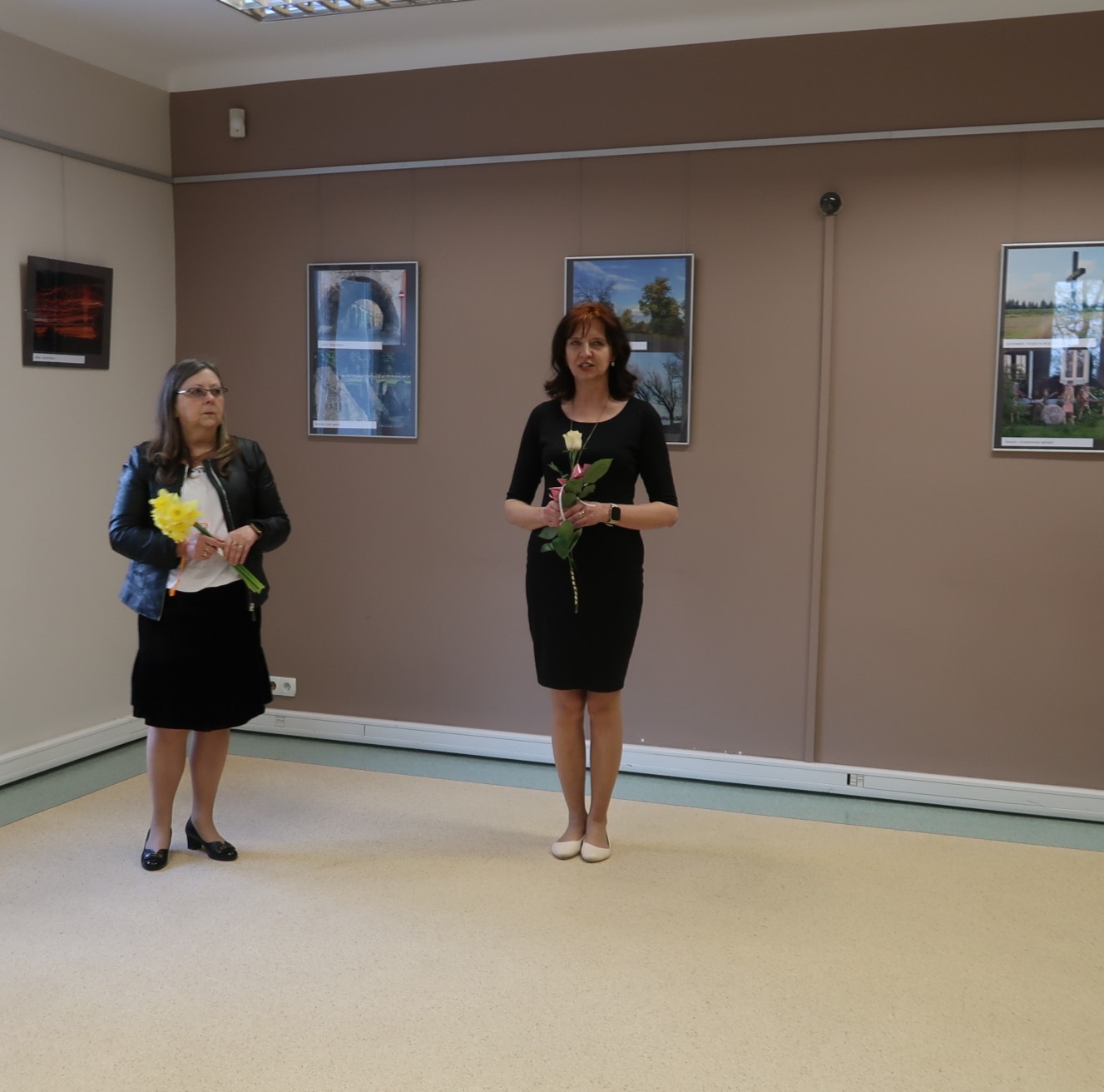 Dwie kobiety z kwiatami w dłoni stoją na środku sali wystawowej.