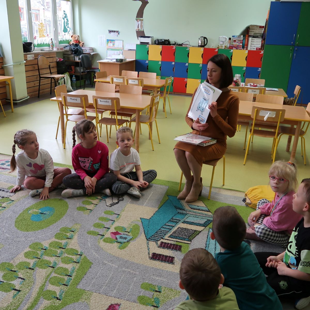 Bibliotekarka siedząc na krześle pokazuje dzieciom kartki z ilustracjami emocji