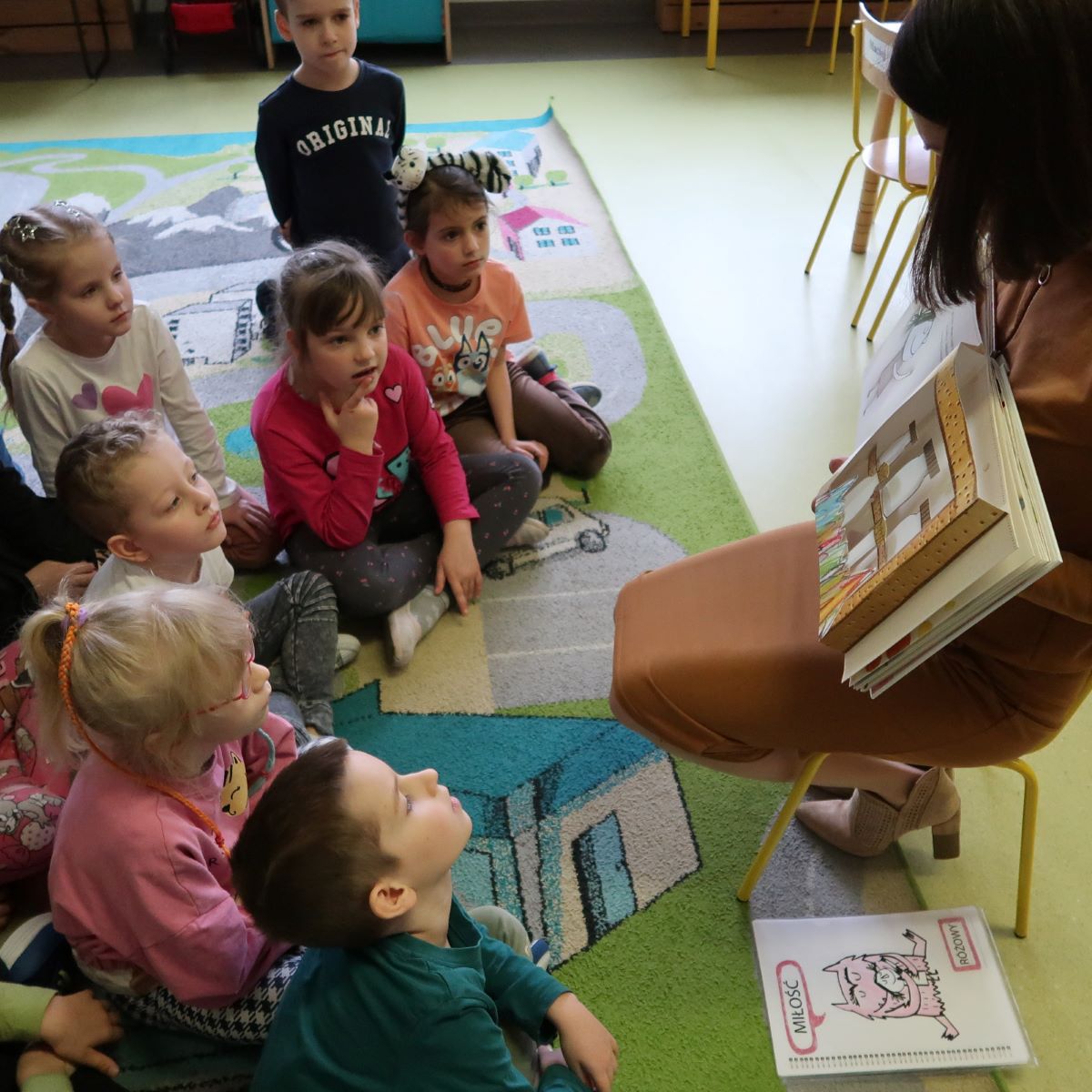 Bibliotekarka siedząc na krześle pokazuje dzieciom książkę z teatrzyku kamoshibai.
