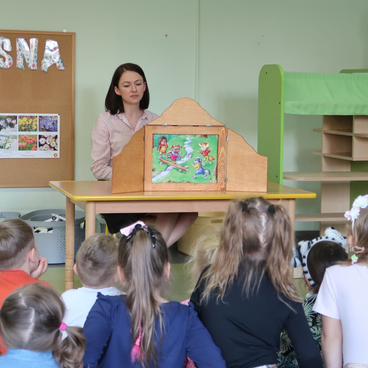 kolorowy-potwor-4 Nauczycielka siedząc za biurkiem przedstawia dzieciom ilustracje teatrzyku kamishibai