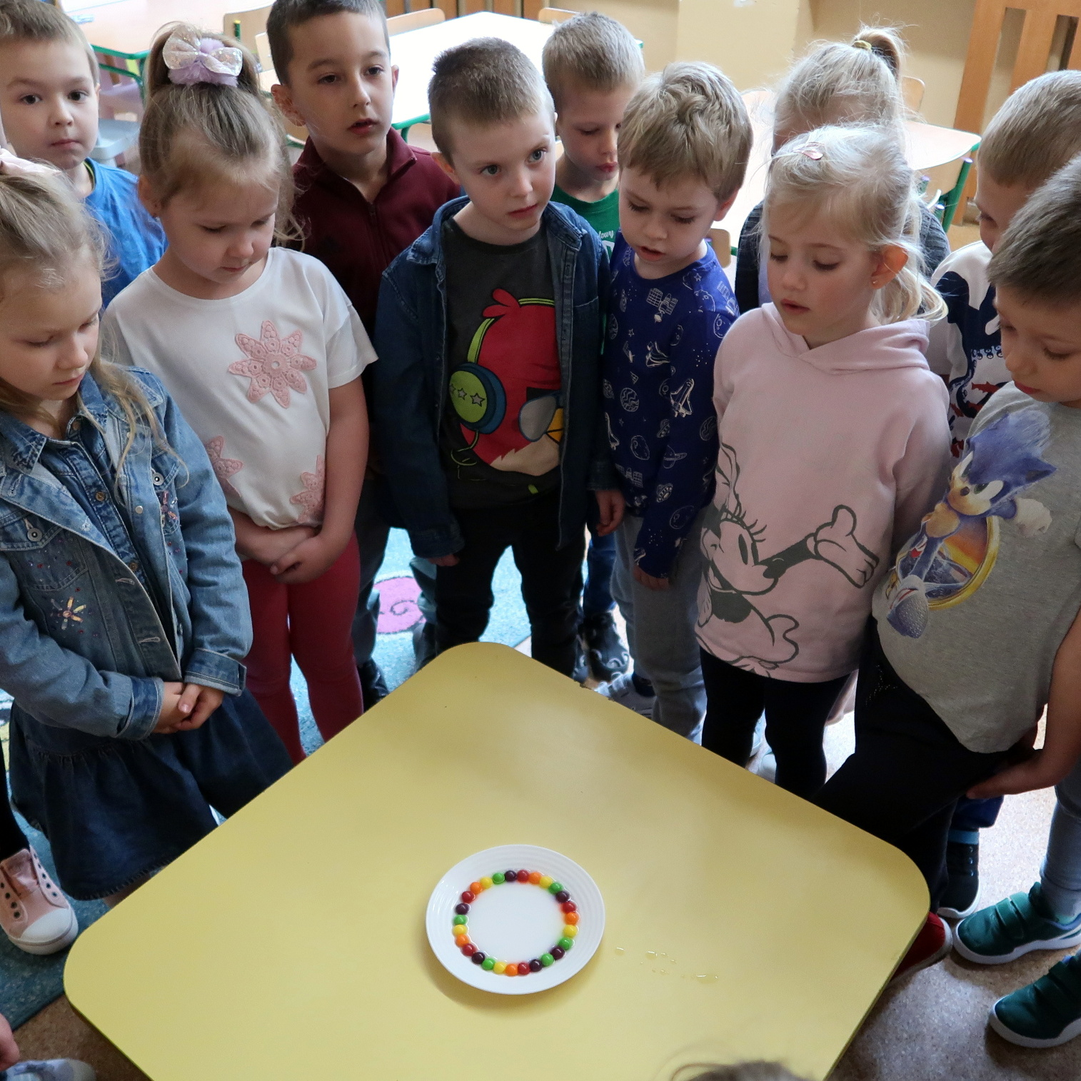 Dzieci stojąc nad stołem obserwują talerzyk z różnokolorowymi kulkami