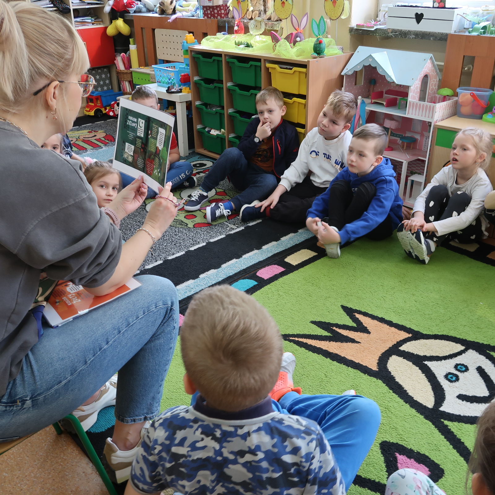 Dzieci słuchają nauczycielki siedząc na dywanie w klasie szkolnej