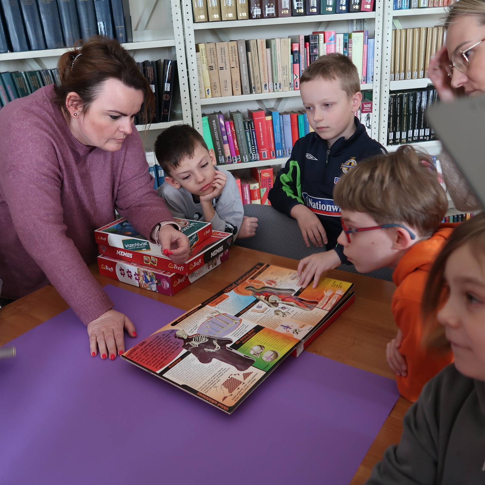 odwiedziny-PSP-12-2 Bibliotekarka pokazuje grupie dzieci ilustrowane książki gromadzone w bibliotece.