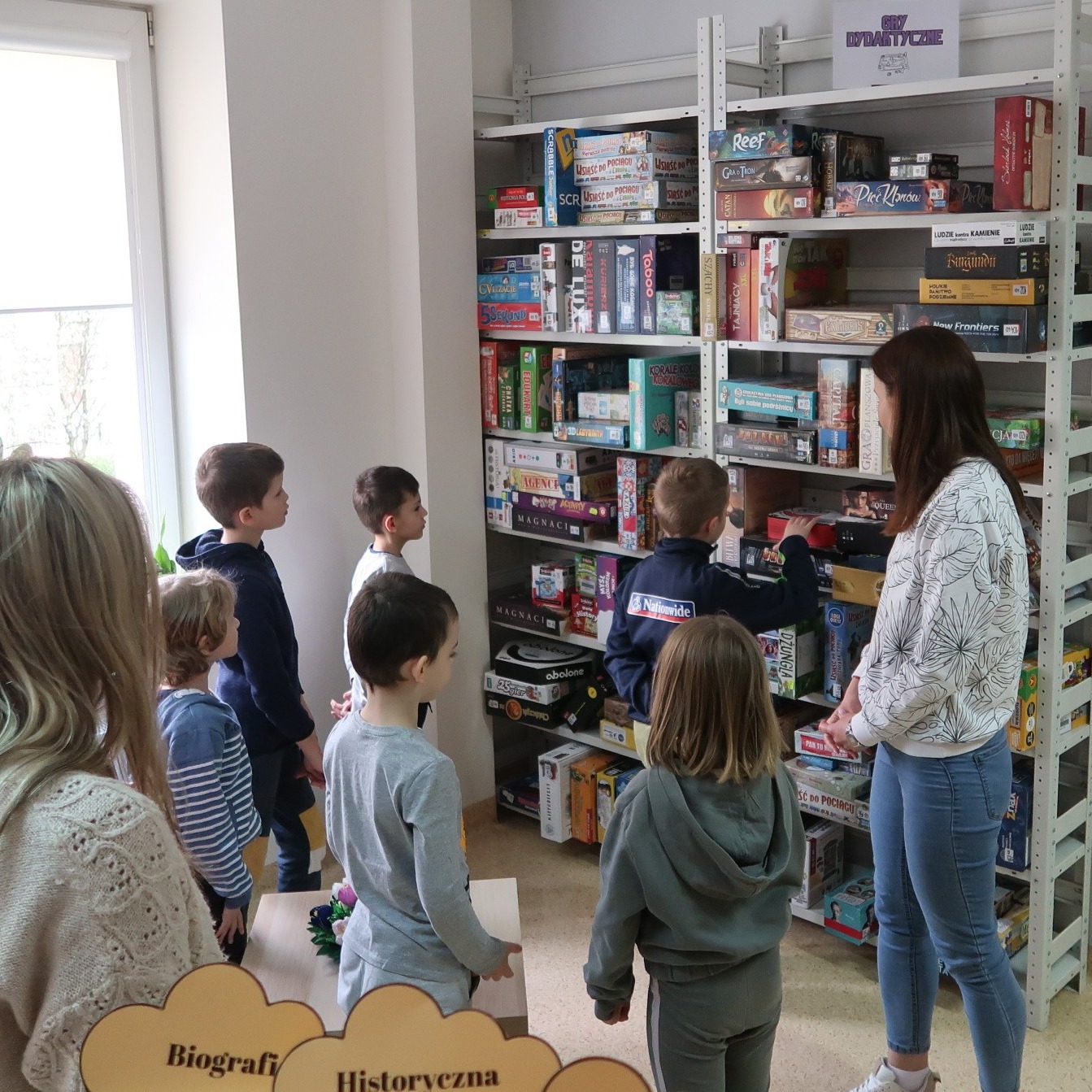 odwiedziny-PSP-12-3 Bibliotekarka pokazuje grupie dzieci gry dydaktyczne zebrane w bibliotece