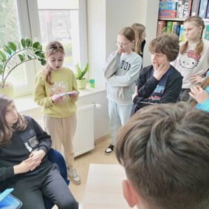 rozGRYzc-biblioteke-1 Uczennica odczytuje zapisaną na kartce papieru zagadkę do rozwiązania pozostali uważnie słuchają