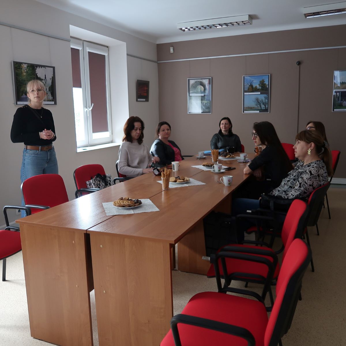 Grupa osób w sali konferencyjnej siedzi wokół stołu i słucha z uwagę prelegentki