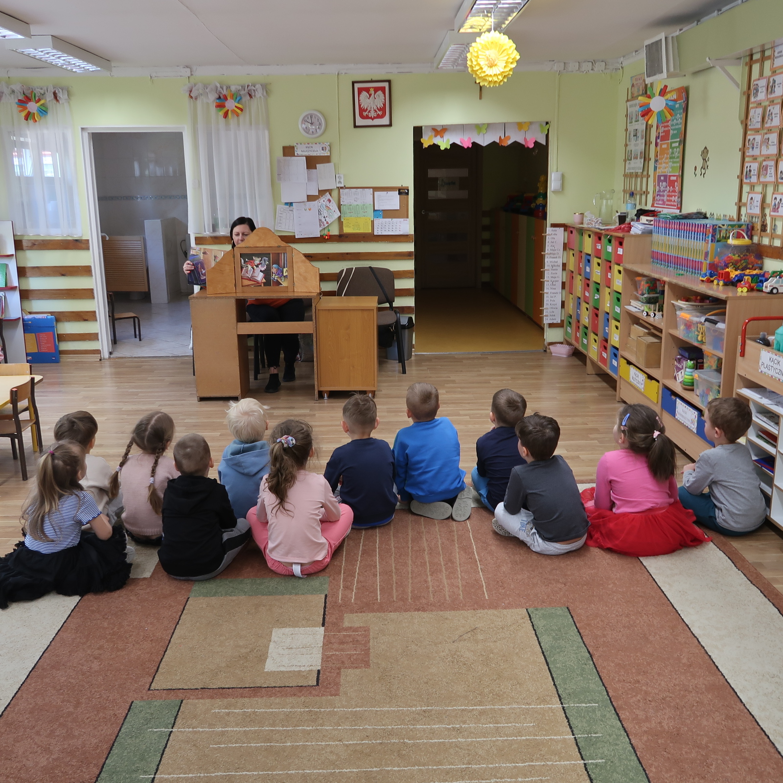 zajecia-PSP-12-2 Dzieci siedząc na dywanie uważnie słuchają opowieści z teatrzyku kamishibai