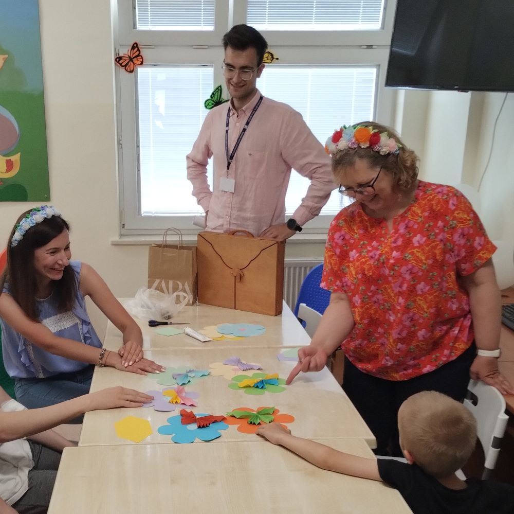 Grupa osób, w tym dwie kobiety i mężczyzna, zbiera się wokół stołu i przeglądają prace plastyczne wykonane z kolorowych kartek papieru