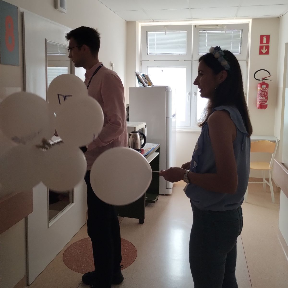 Dwie osoby stoją przed drzwiami prowadzącymi do pokoju szpitalnego w ręku trzymają balony.