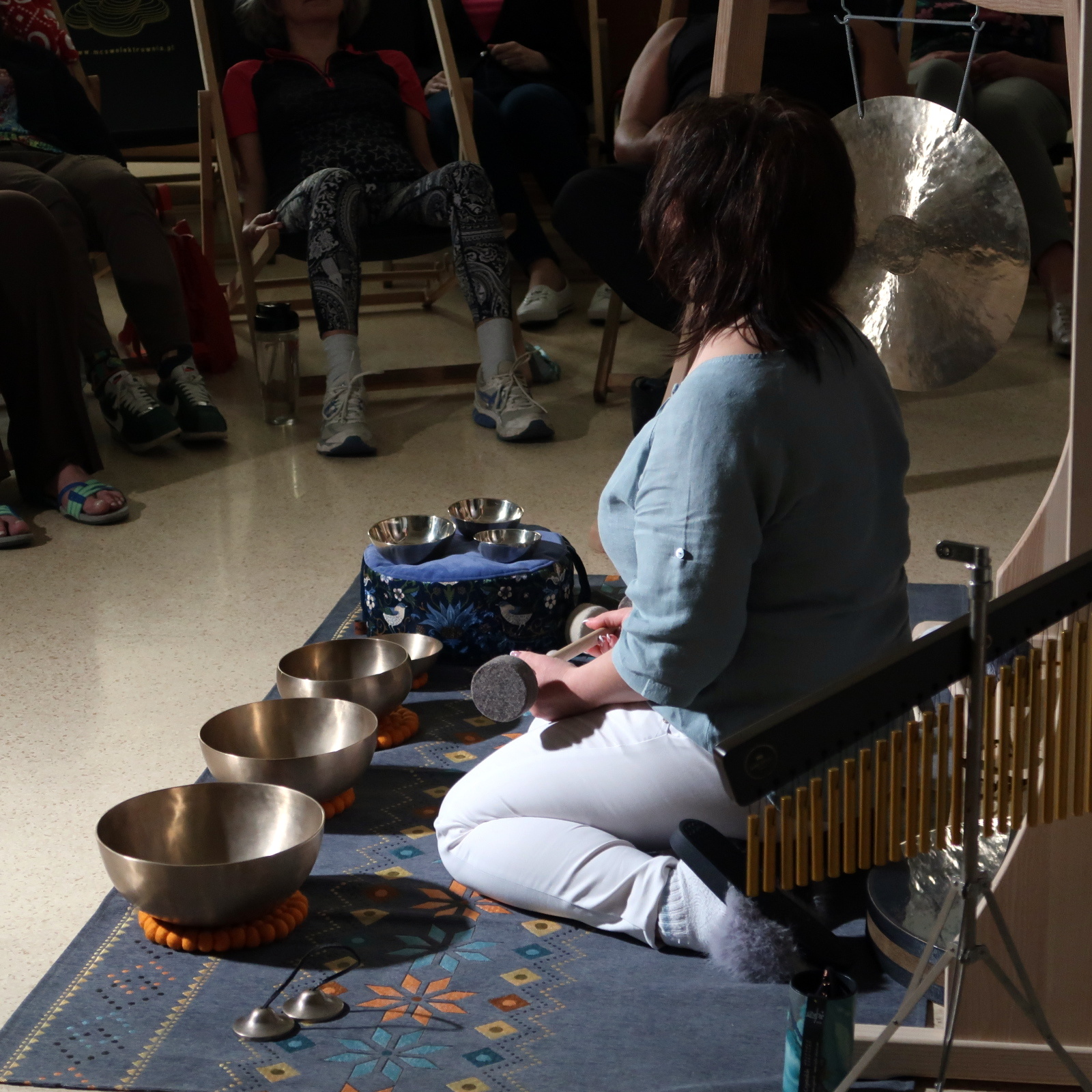 Kobieta klęczy na dywanie z pałką w dłoniach, przed sobą ma ustawione w rzędzie misy różnej wielkości.