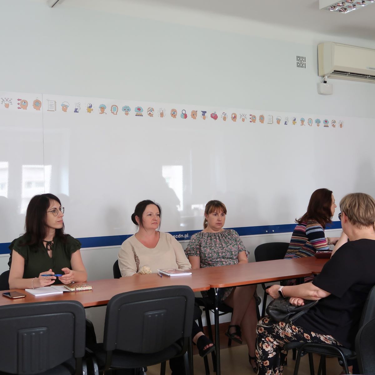 Grupa kobiet siedzących przy długim stole w sali konferencyjnej. Są zajęci rozmową i robieniem notatek.