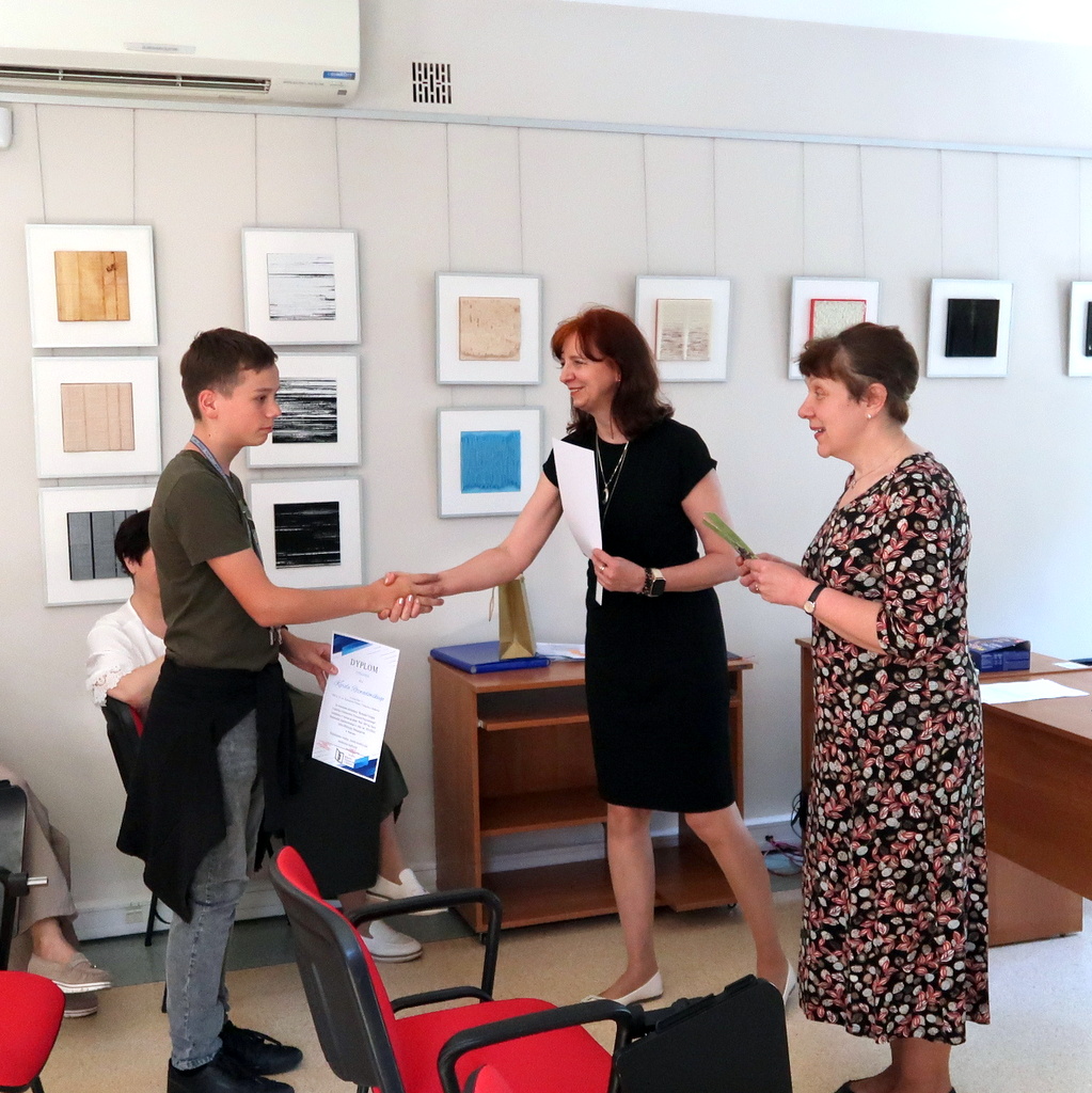 W pokoju, w którym na ścianach wiszą oprawione prace plastyczne, młody chłopak otrzymuje dyplom od kobiety w czarnej sukience, podczas gdy inna kobieta w kwiecistej sukience przygląda się i wręcza kolejny dokument.