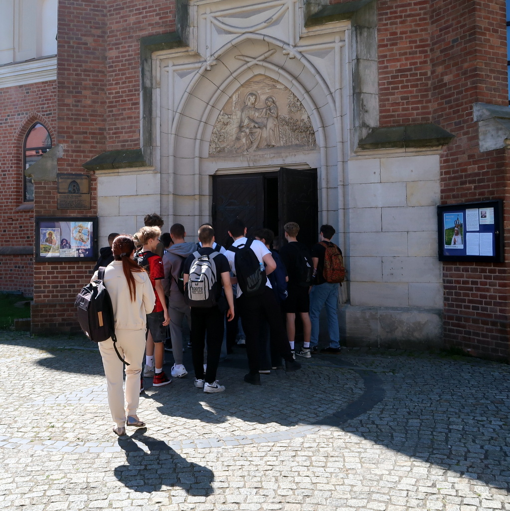 Grupa ludzi stoi przy wejściu do budynku świątyni z czerwonej cegły i zdobionym kamiennym łukiem.