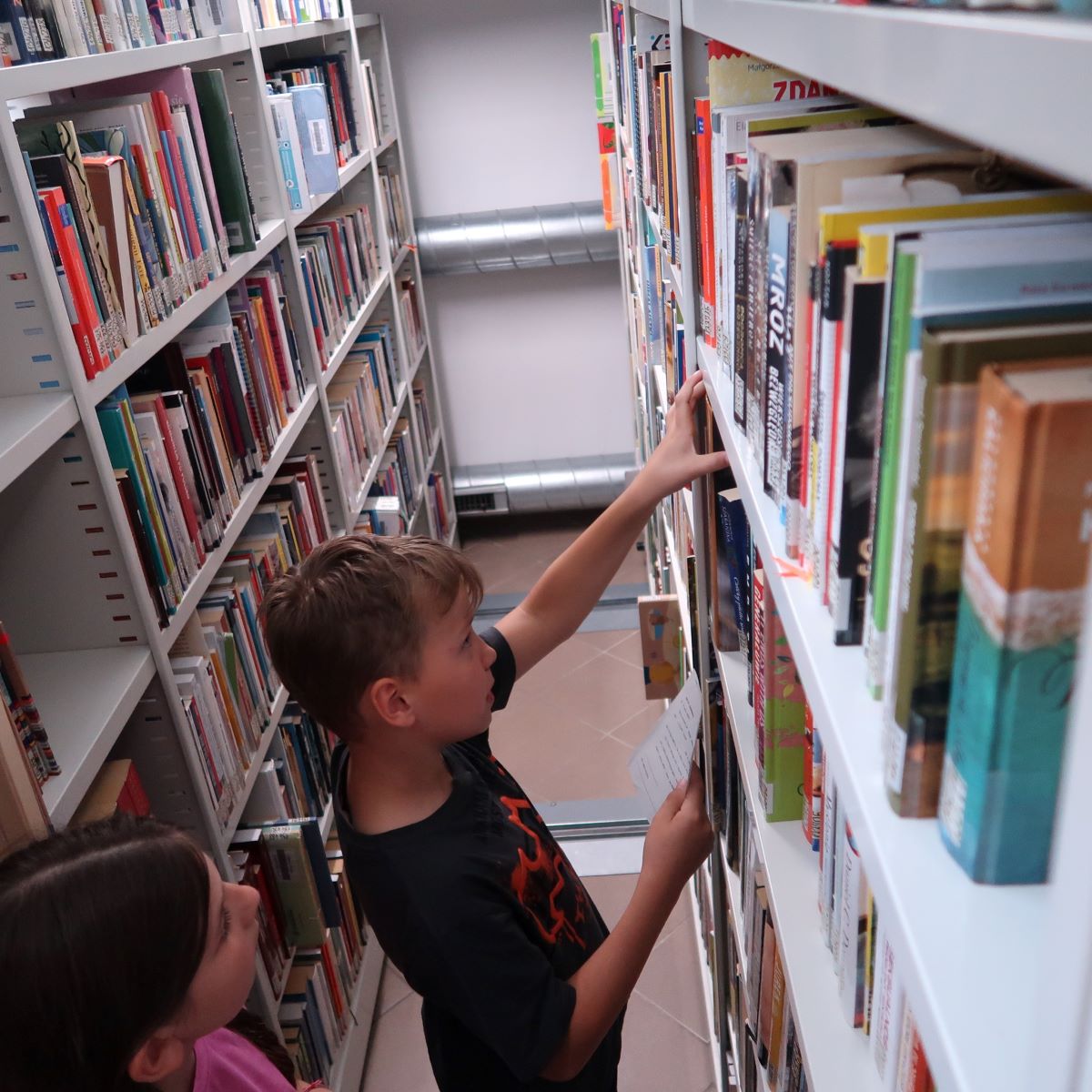 Chłopiec sięga po książkę stojącą na wysokiej półce regału bibliotecznego.