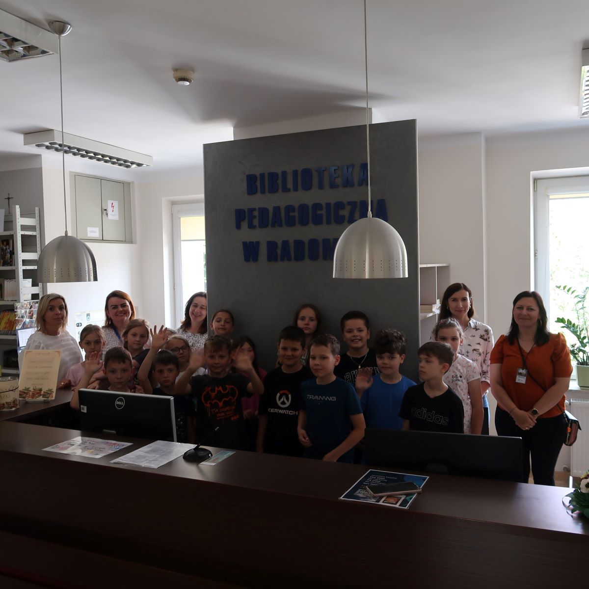 Grupa dzieci i dorosłych pozuje do zdjęcia stojąc w bibliotece za ladą z komputerami. Nad nimi widniał napis: „Biblioteka Pedagogiczna w Radomiu”.