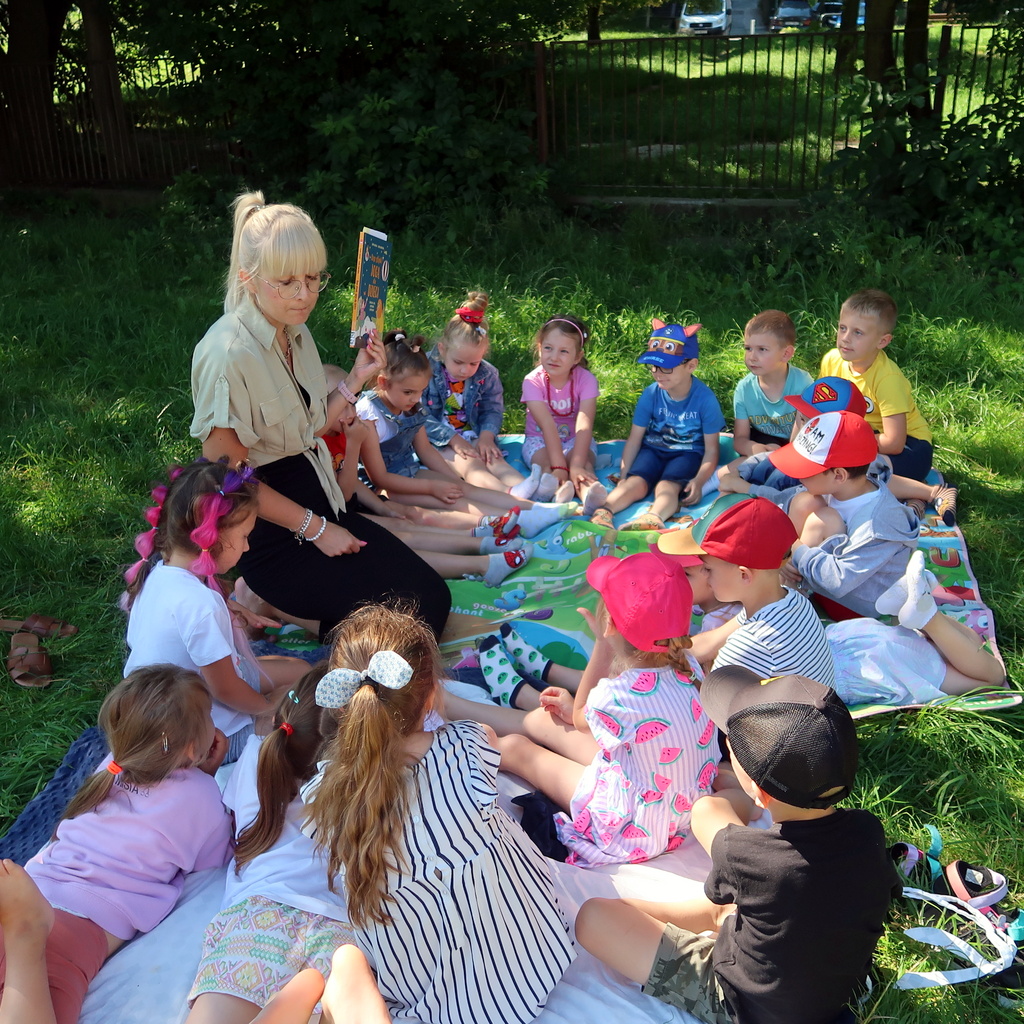 Dzieci siedząc na trawie słuchają pani prowadzącej zajęcia czytelnicze