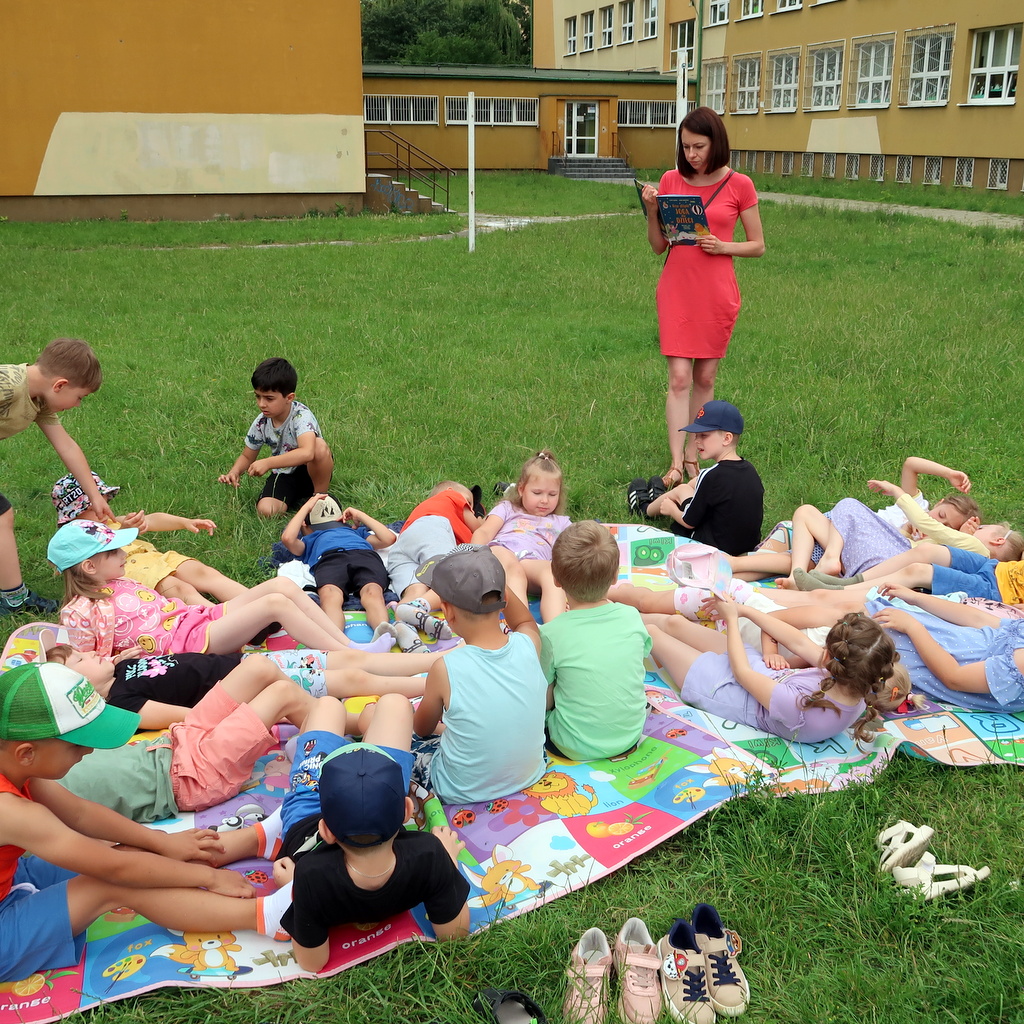 Dzieci siedząc na trawie boiska szkolnego słuchają bajki czytanej przez nauczycielkę