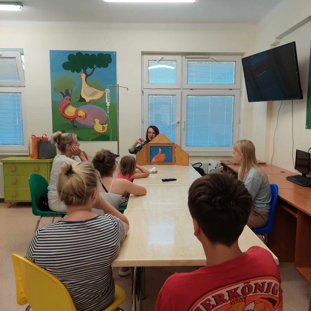 Kobieta siedząca na końcu stołu konferencyjnego prezentuje dzieciom teatrzyk kamishibai.