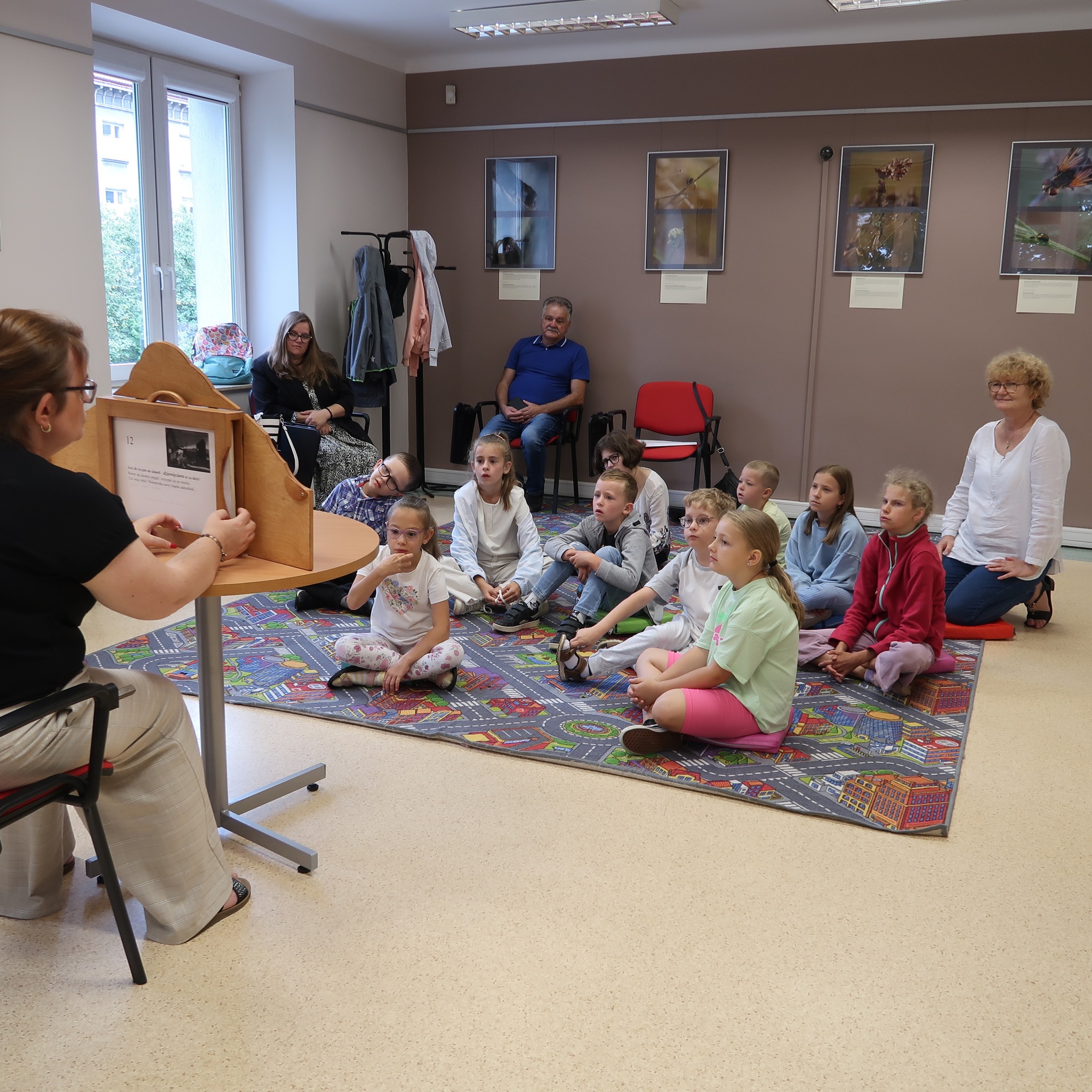 Dzieci siedząc na dywanie w sali dydaktycznej oglądają teatrzyk kamishibai.