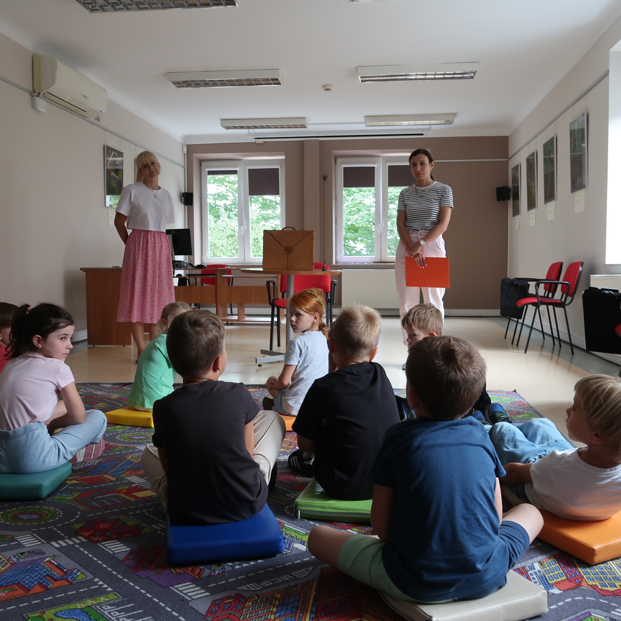 Dzieci siedząc na dywanie słuchają pań stojących przed nimi