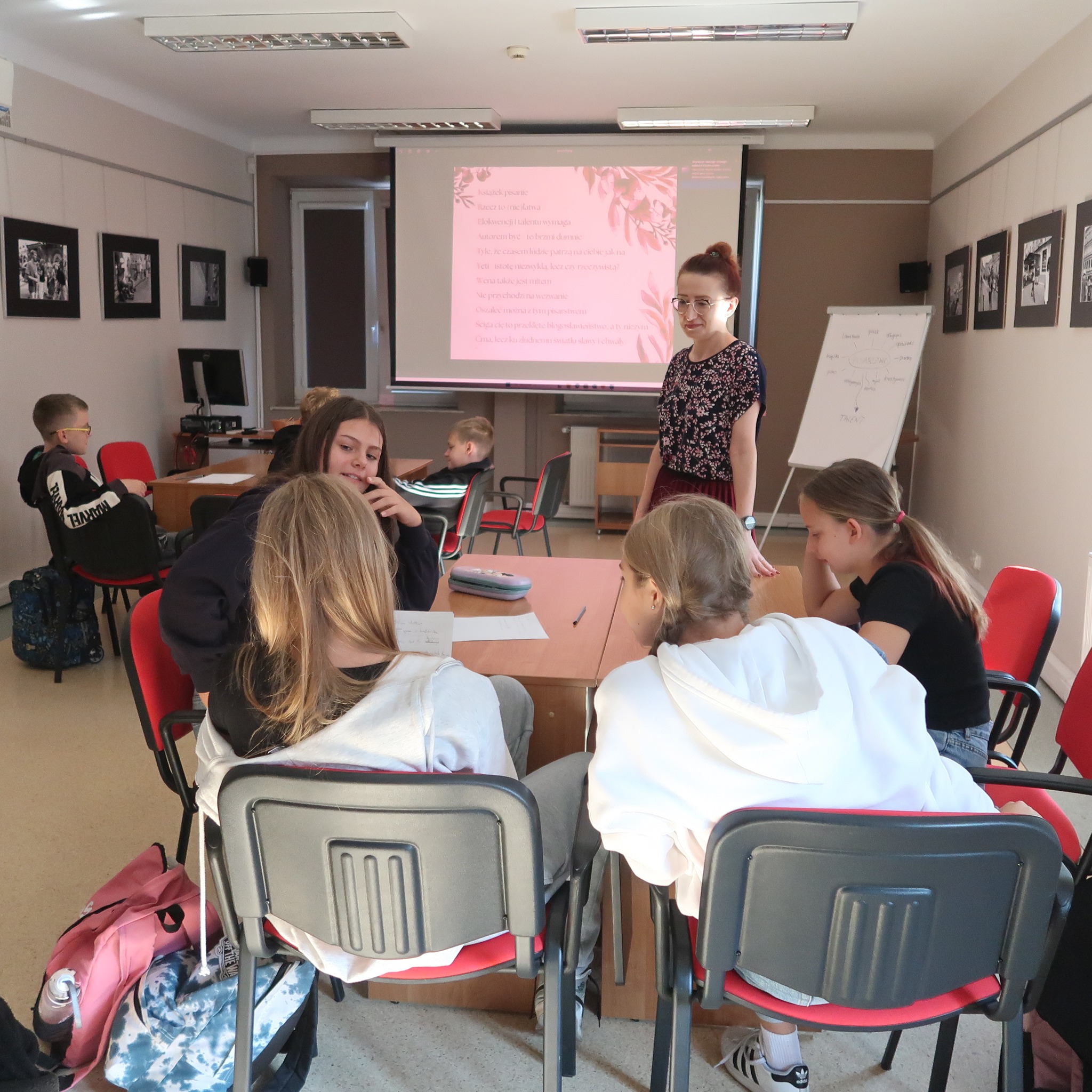 Cztery dziewczynki siedząc przy biurkach słuchają stojącej pani.