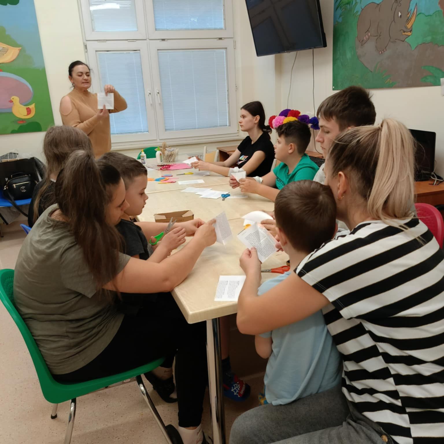 Dzieci siedząc przy stole wykonują prace plastyczne pod kierunkiem prowadzącej zajęcia