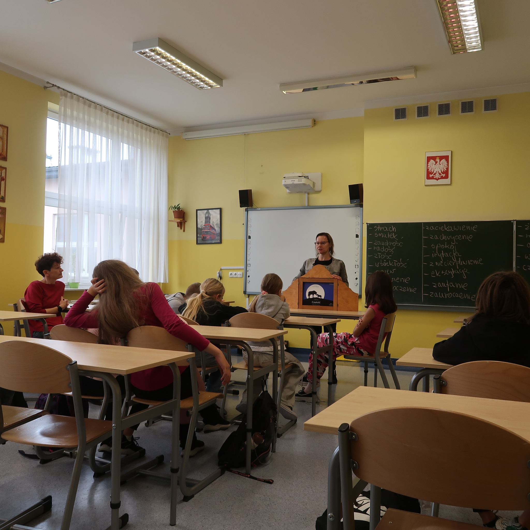 Zajęcia w klasie szkolnej. Pani stojąc przodem do klasy prowadzi lekcję, prezentuje planszę z teatrzyku kamishibai.